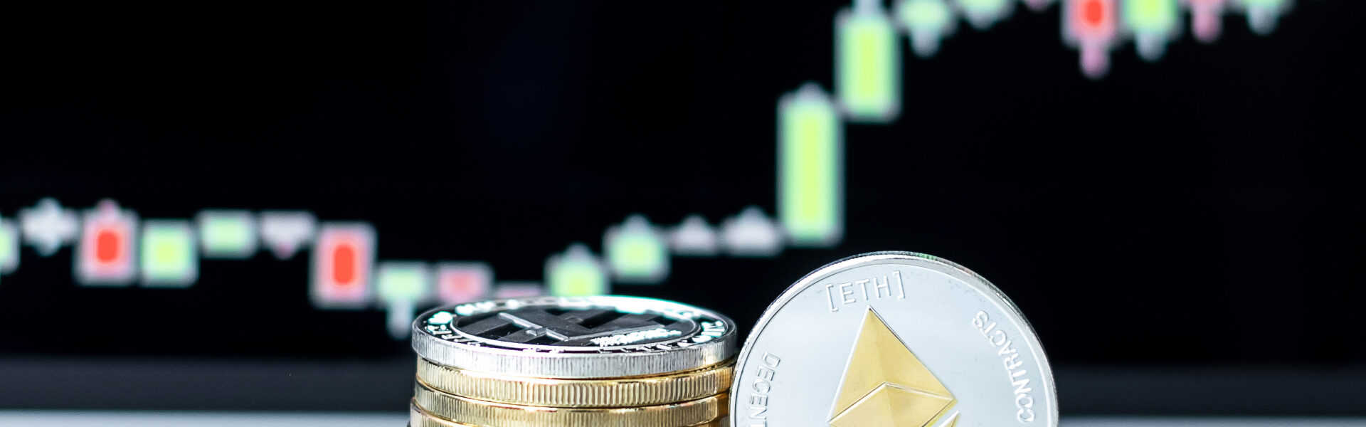 A close-up of stacked coins with a prominent silver Ethereum (ETH) token in focus, placed in front of a glowing candlestick chart showing market volatility and potential recovery trends.